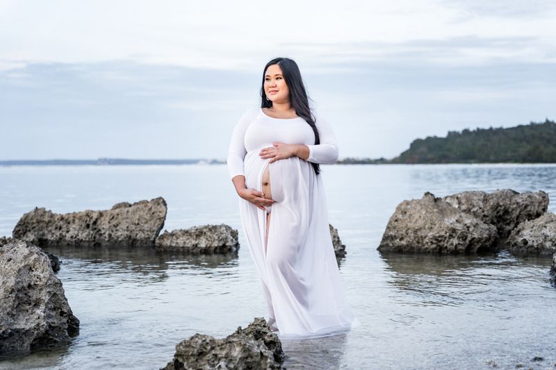 maternity photoshoot woman in white long dress holding her pregnant belly with ocean on background